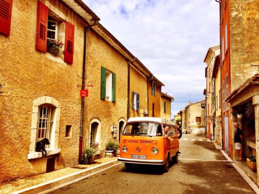 Iconic VW orange van in a narrow street through historical buildings. 