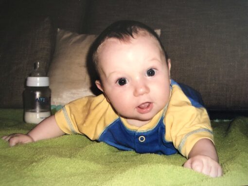 Baby crawling looking straight into camera with milk bottle behind him.