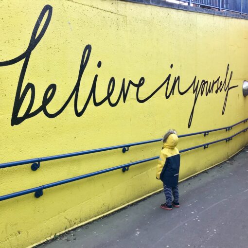 Kid looking up scribble 'Believe in yourself' in black over a yellow wall.