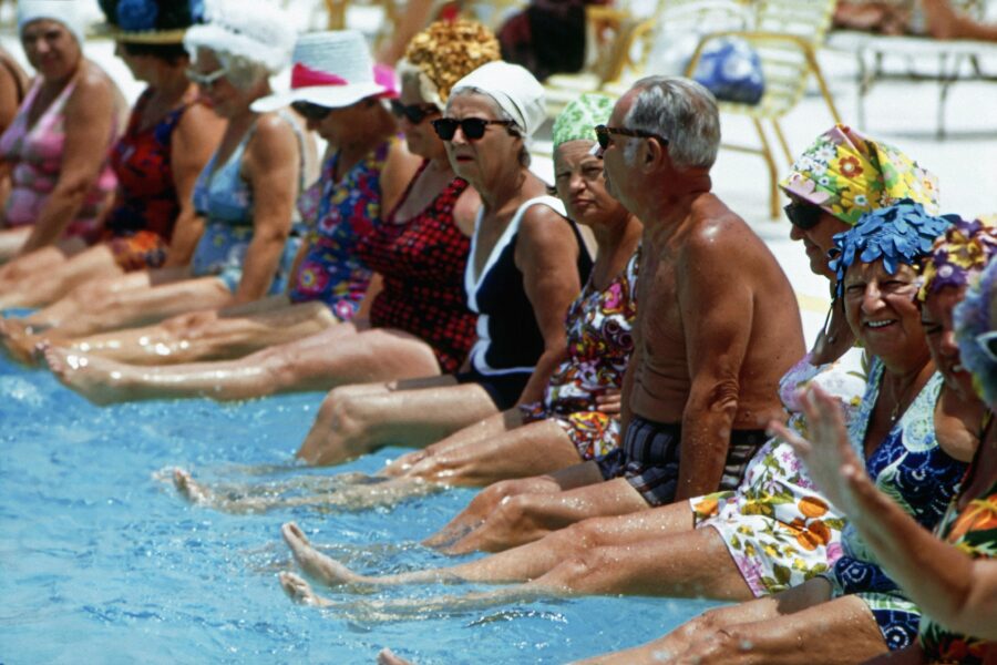 Elderly folks in swimwear sitting along the pool rim and kicking their feet in the water.