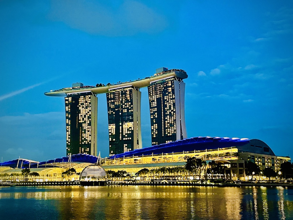 The iconic Singapore Marina Bay Sands with 3 towers linked by a 'boat' structure are the top, with lighted buildings below.