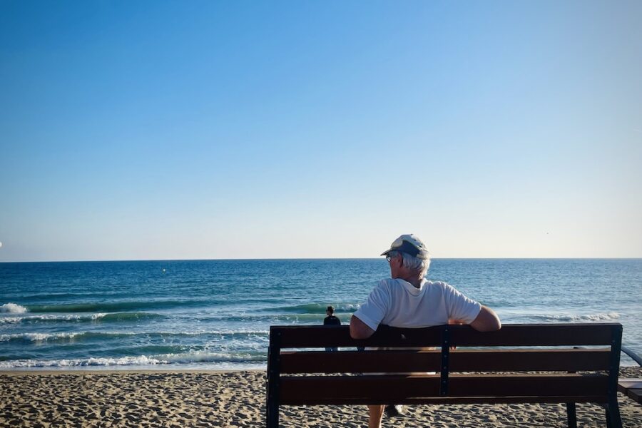 A lone elderly man sits on a bench watching the tides rolling onto the beach.