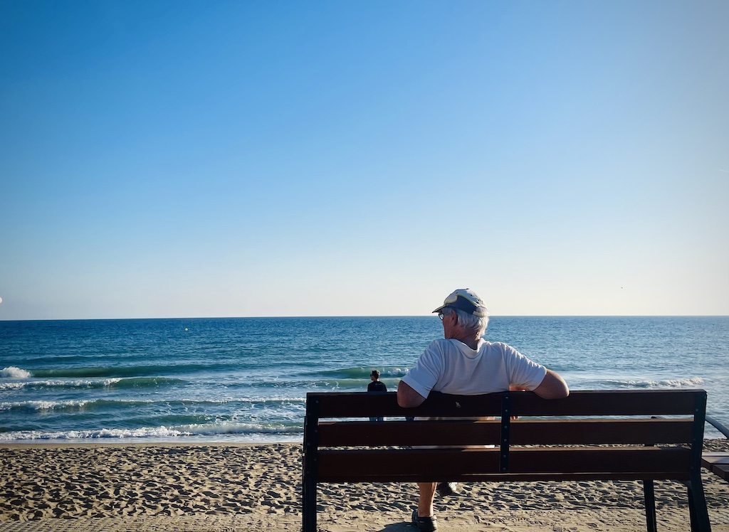 A lone elderly man sits on a bench watching the tides rolling onto the beach.