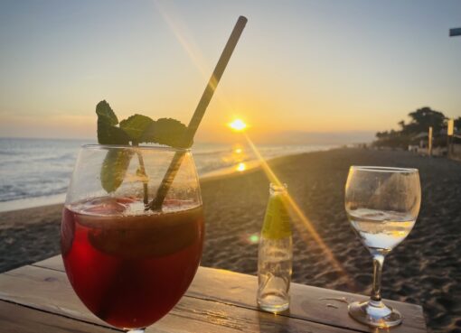 2 glasses of cocktail drinks at beachfront table facing golden sunset.