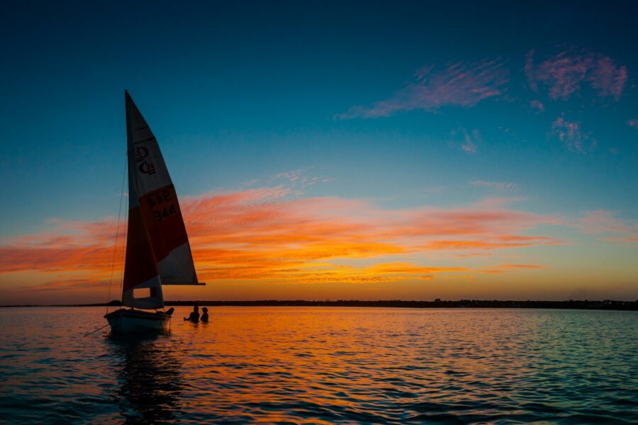 Silhouette of 2 people sitting in shallow ocean water next to their sailboat chatting at sunset.