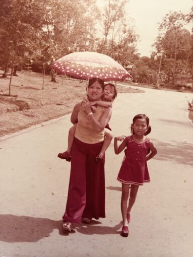 Old photo of a young mother carry 1 child piggy-back holding an umbrella and another daughter walking beside.