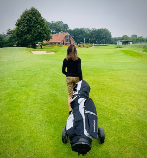 Woman golfer walking towards putting green pulling a golf trolley behind her. 