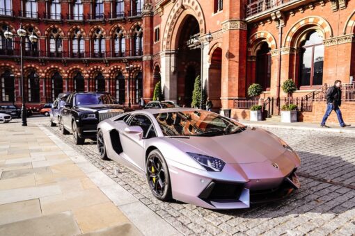 Luxury grey sports car parked in front of a Rolls Royce on a posh street.