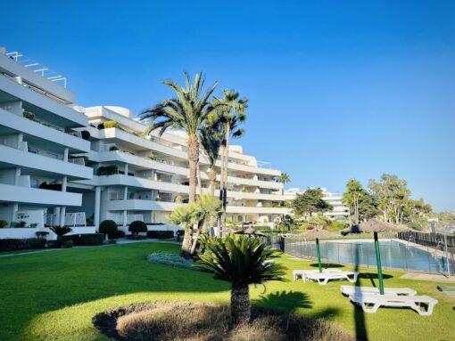 Condominium with a fenced-in pool with sundeck chairs on a green lawn, against clear blue sky.