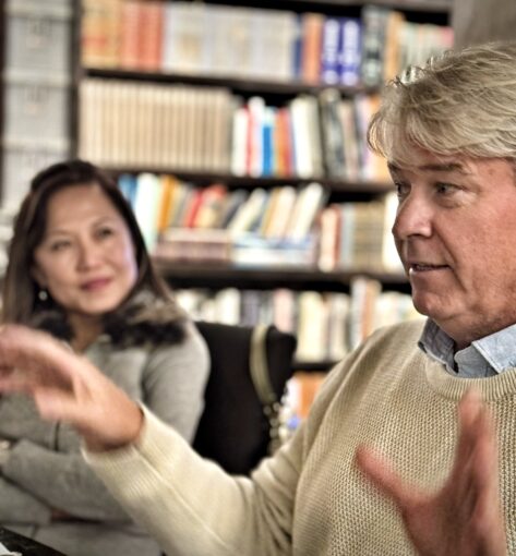 Man gesturing with both hands as he speaks with a women looking at him at the side in a library setting.