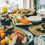 Table with spread of sushi, platter of cut fruit, dips, small bites amidst champagne glasses and dinner plates.